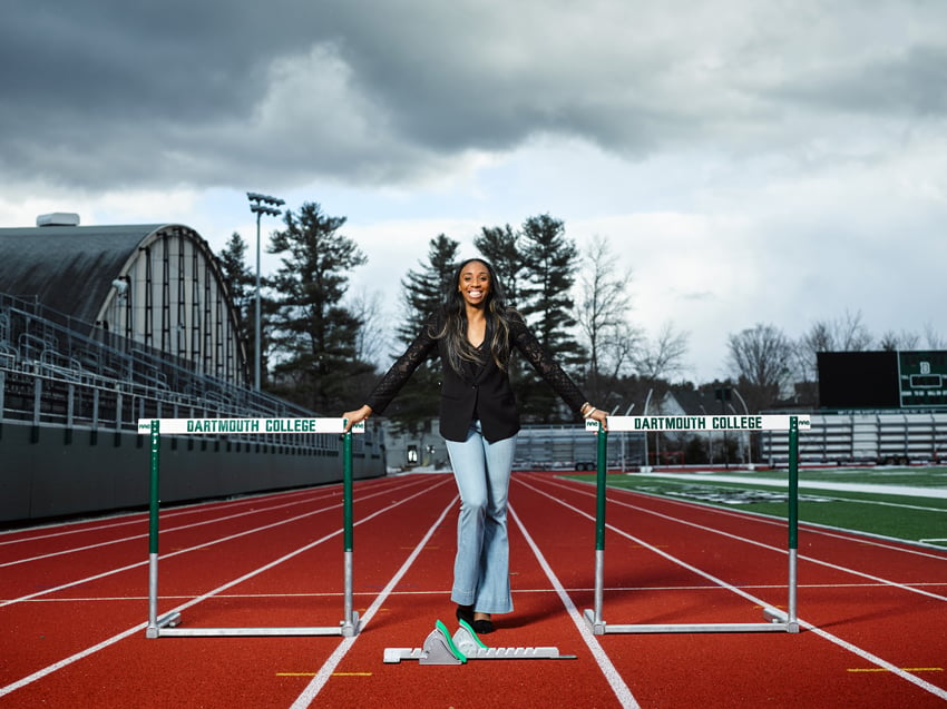 Photographer Doug Levy's photo for Financial Times of Olympian Lashinda Demus. The photo is a portrait of Lashinda wearing jeans and a black blazer posed between two green and white hurdles that say "Dartmouth College" on a race track. 